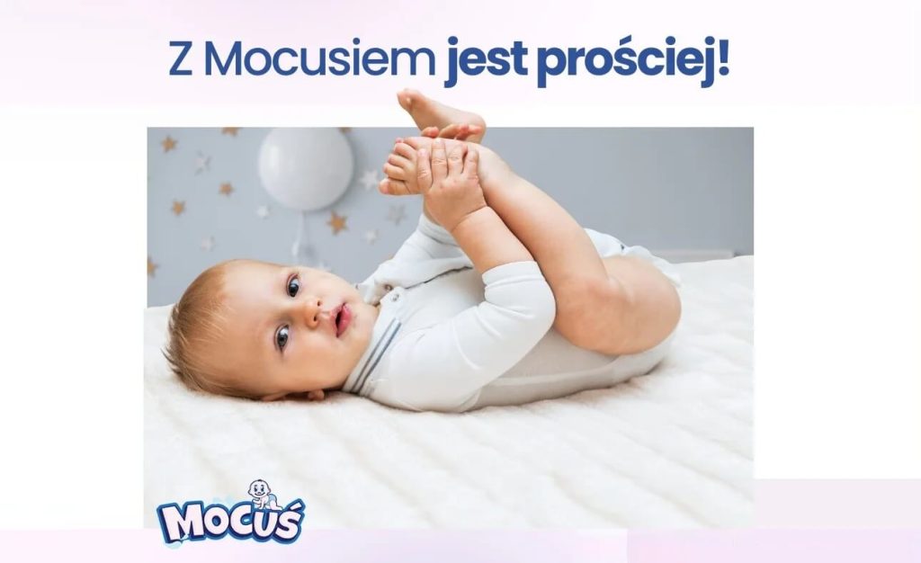 Na zdjęciu widzimy niemowlę leżące na miękkim, białym podłożu w radosnej pozie — z uniesionymi nóżkami i rączkami trzymającymi stopy. Dziecko ubrane jest w białe body i patrzy prosto w obiektyw. Tło ozdobione jest gwiazdkami i balonem. Na górze widnieje napis „Z Mociusiem jest prościej!”, a w rogu znajduje się logo z nazwą „Mocuś”.