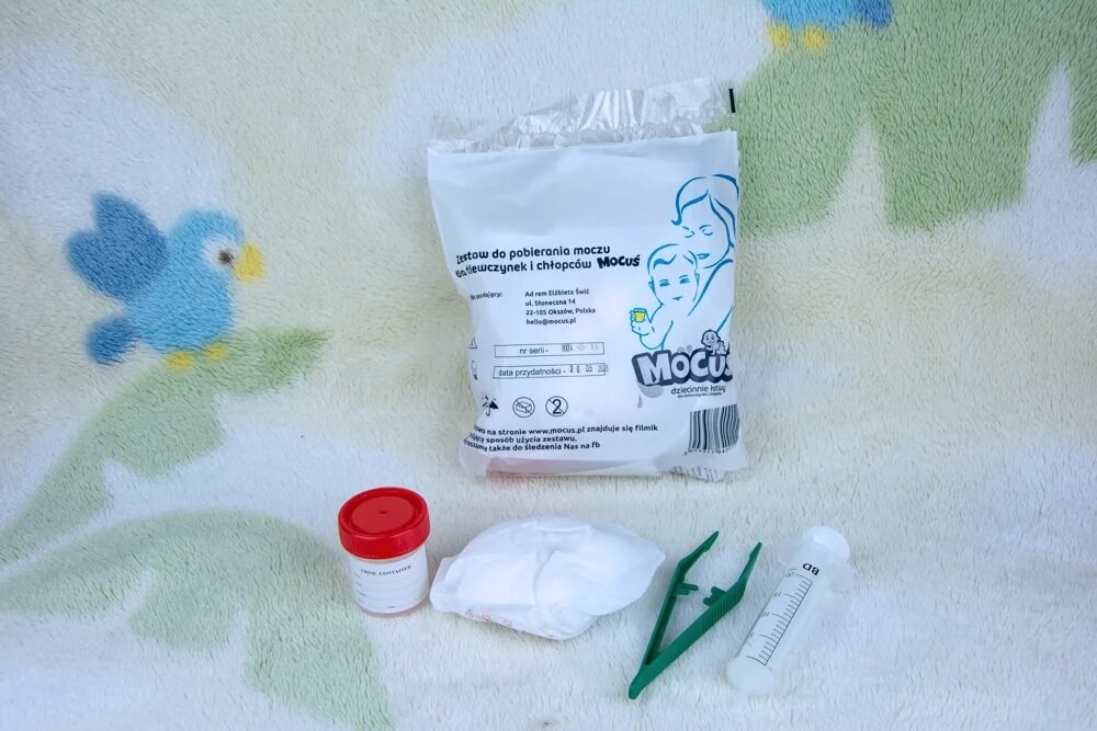 Na zdjęciu znajduje się zestaw medyczny do pobierania moczu u niemowląt dla dziewczynek i chłopców. W skład kompletu wchodzi pojemnik z czerwonym korkiem, chusteczka higieniczna, strzykawka oraz zielony uchwyt. Całość spoczywa na miękkim materiale z dziecięcym wzorem ptaków i liści, co nadaje scenie domowy, troskliwy charakter. Opakowanie zawiera szczegółowe informacje w języku polskim, w tym datę ważności i oznaczenia zgodności z normami medycznymi.